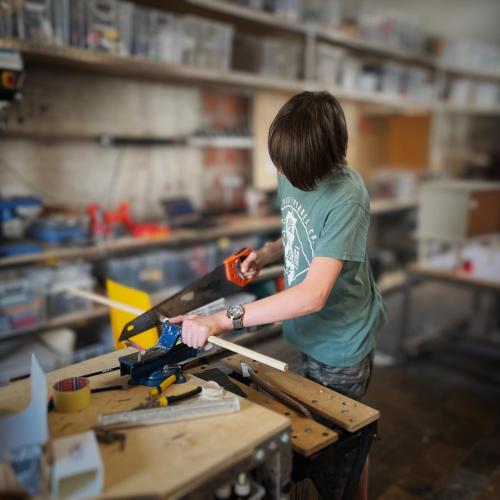 In de workshopruimte is een jonge deelnemer naarstig aan het werk. Met een handzaag korten ze een houten pin af tot op de juiste lengte.
