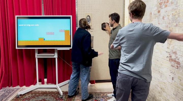 Begeleider Frederik test samen met twee deelnemers een van de puzzels uit. Op een groot scherm zie je een videogame. De spelers raken tegelijkertijd verschillende objecten in de ruimte aan.